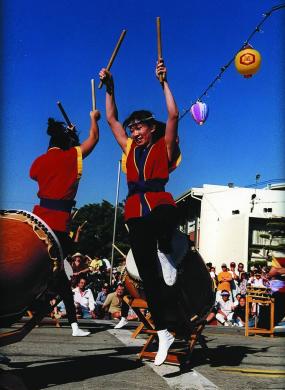 Two people raise their arms holding drums sticks dressed in traditional taiko garb